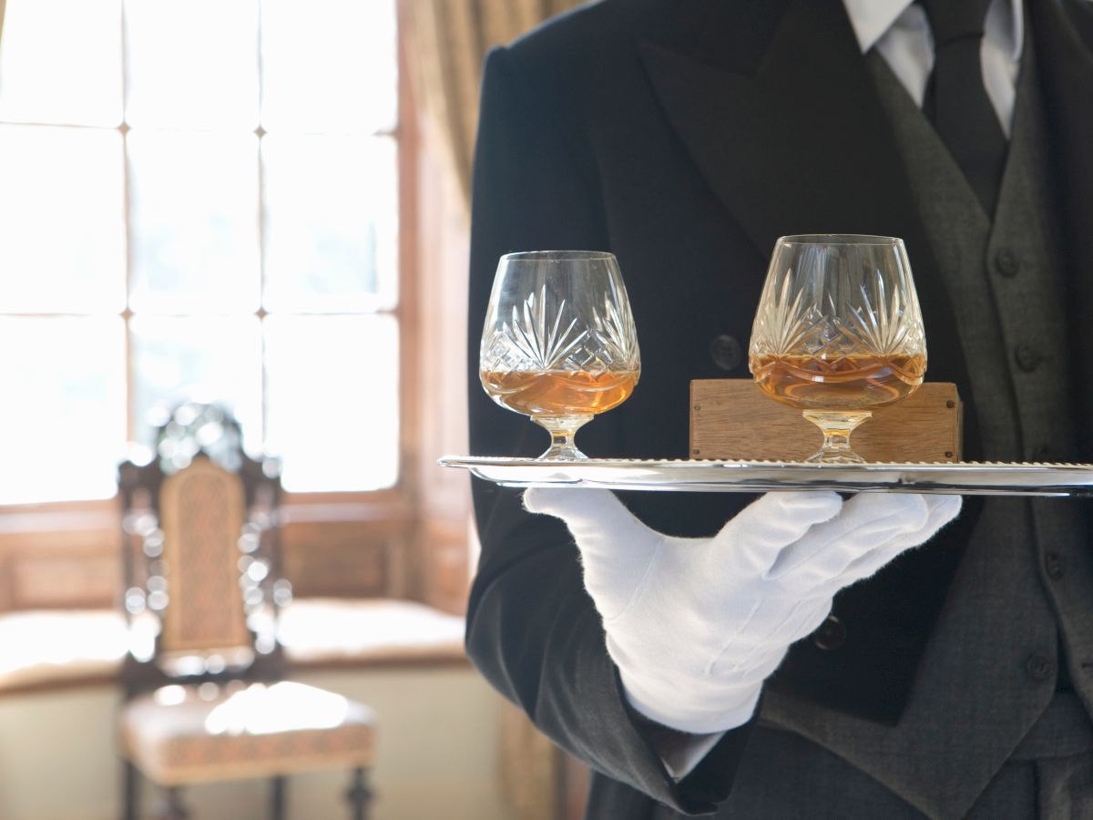 A person in formal attire holds a tray with two glasses of amber liquid, standing in an elegantly furnished room.
