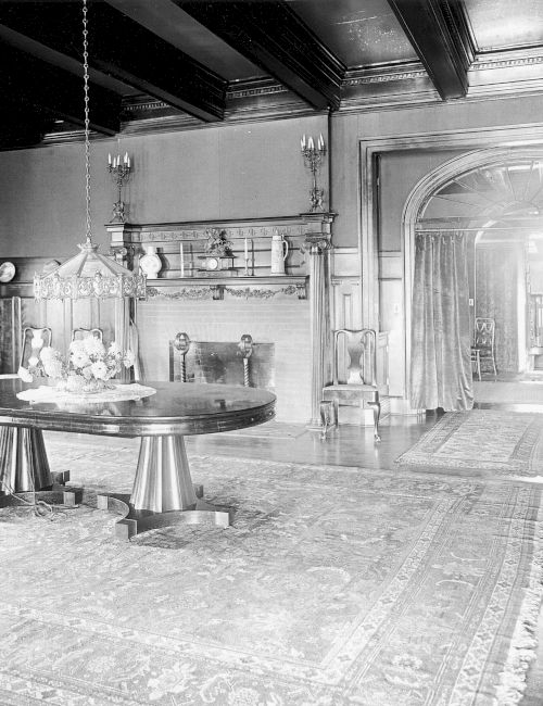 A large, elegant room features a wooden table, ornate rugs, a fireplace, and an arched doorway with drapes, in a classic interior style.