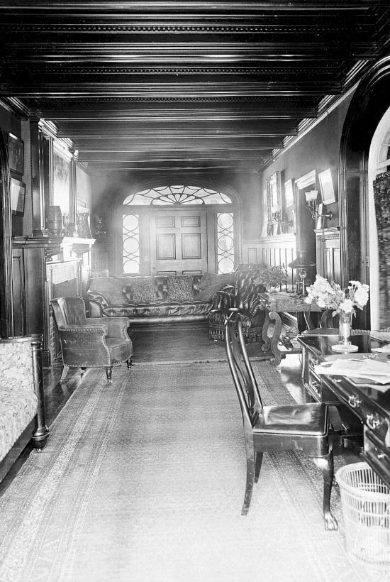 A vintage room with ornate furniture, including sofas, chairs, tables, and framed pictures on wood-paneled walls.