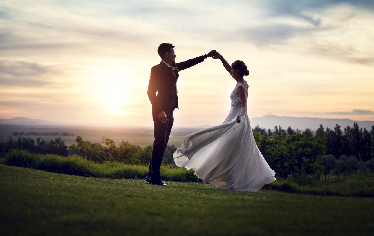 A couple dances elegantly on a grassy field at sunset, with the sky painted in soft colors and trees in the background, creating a romantic scene.