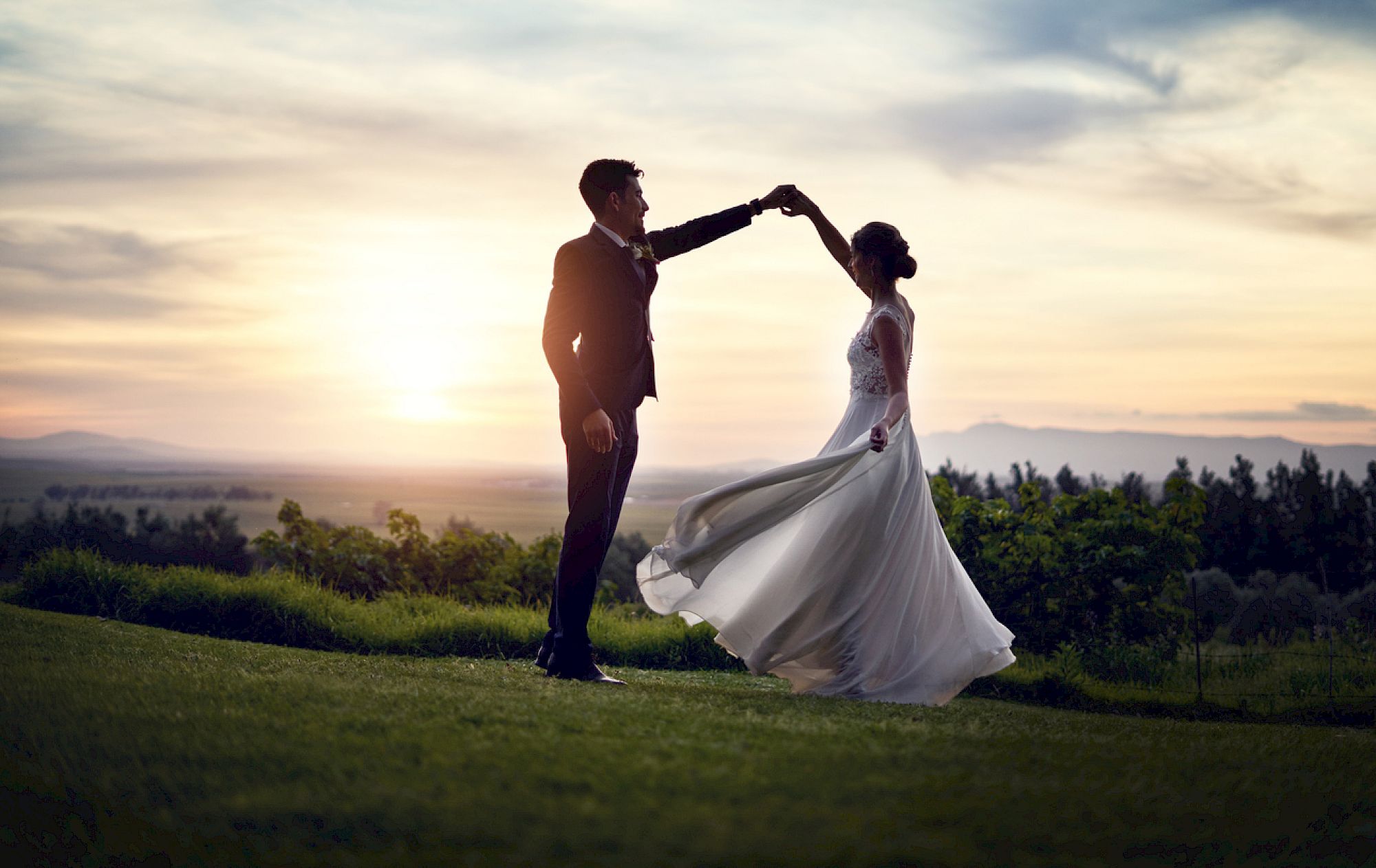 A couple dances elegantly on a grassy field at sunset, with the sky painted in soft colors and trees in the background, creating a romantic scene.