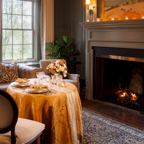A cozy dining room with a set table, candles and flowers, and a roaring fireplace in the background.
