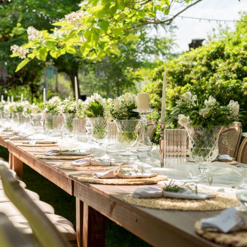 A long outdoor table set for an event with elegant tableware, candles, and floral arrangements amidst a lush garden backdrop.