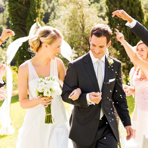 A couple is walking down an aisle at a wedding ceremony, with guests celebrating and throwing confetti outdoors.