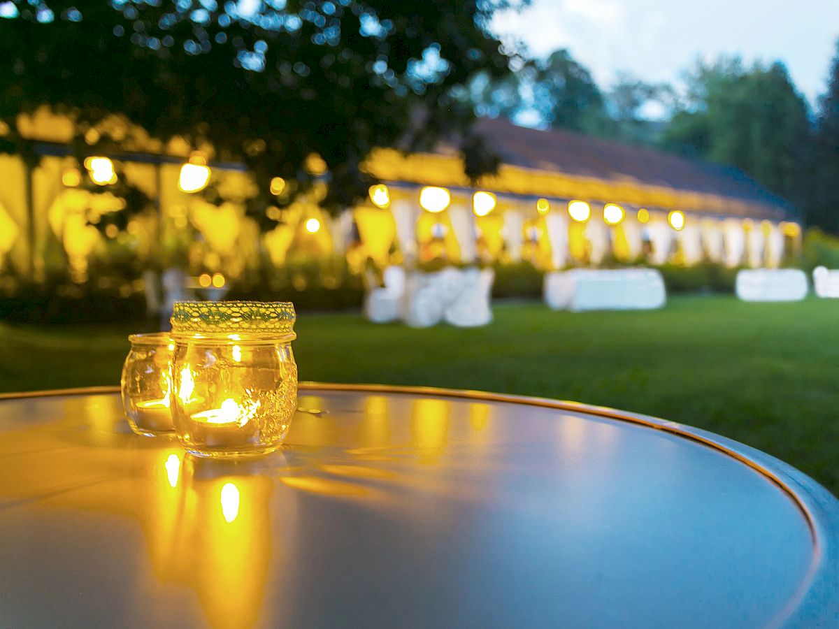 A candlelit jar on a table, with a softly lit outdoor venue in the background, possibly prepared for an evening event.