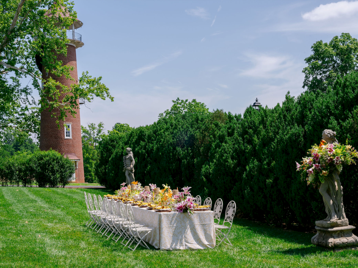 Outdoor garden wedding setup: leafy trees, a brick tower, long table with flowers and chairs, plus statues under blue sky.