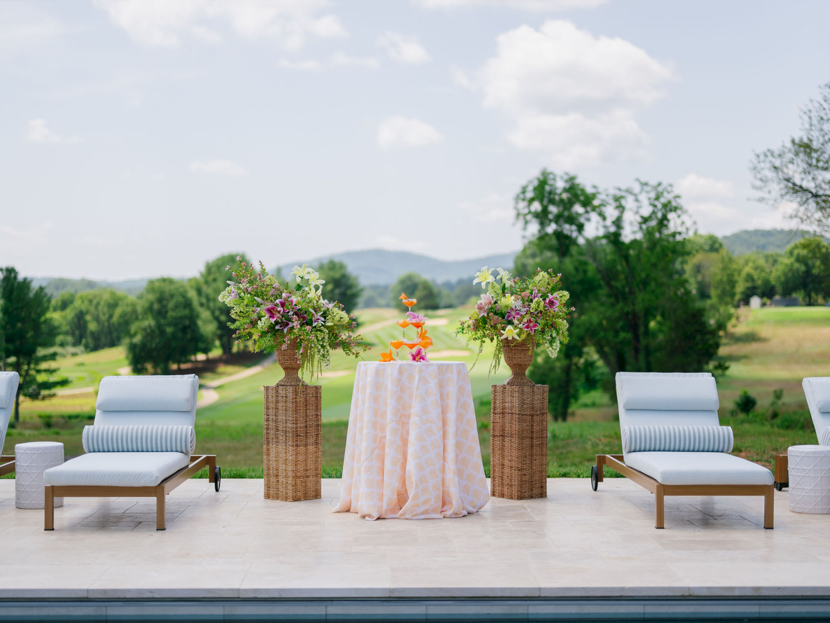 Outdoor wedding setup: white chairs, a covered table with bright flower arrangements, and a lush countryside backdrop.