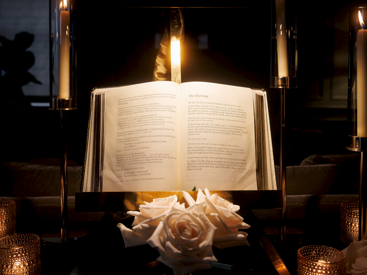 Open Bible on a stand at a candlelit wedding, with rings, white flowers, and tall lit candles behind it.