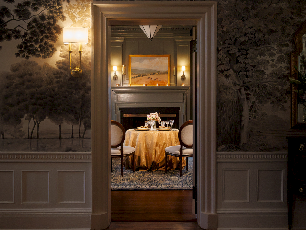 An elegant dining room is visible through an open doorway, with a set table, chairs, and warm lighting.