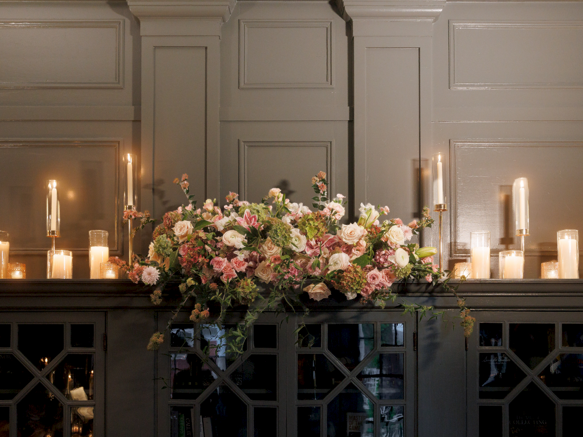 A large floral arrangement sits on a church altar, surrounded by tall lit candles and soft warm light.