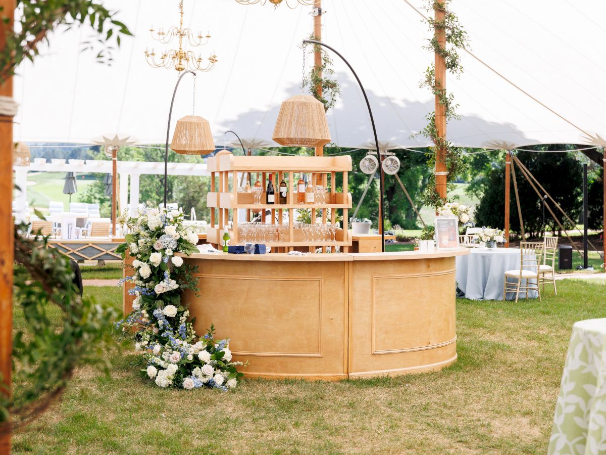 An outdoor wedding drink bar with hanging pendant lights, bottles, and floral decor under a tent canopy.