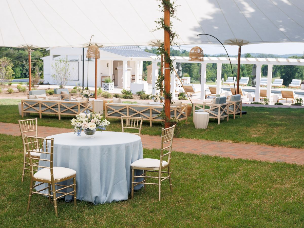 Outdoor wedding setup with white tent, rows of chairs and round table, floral centerpieces, and wooden arch accents.