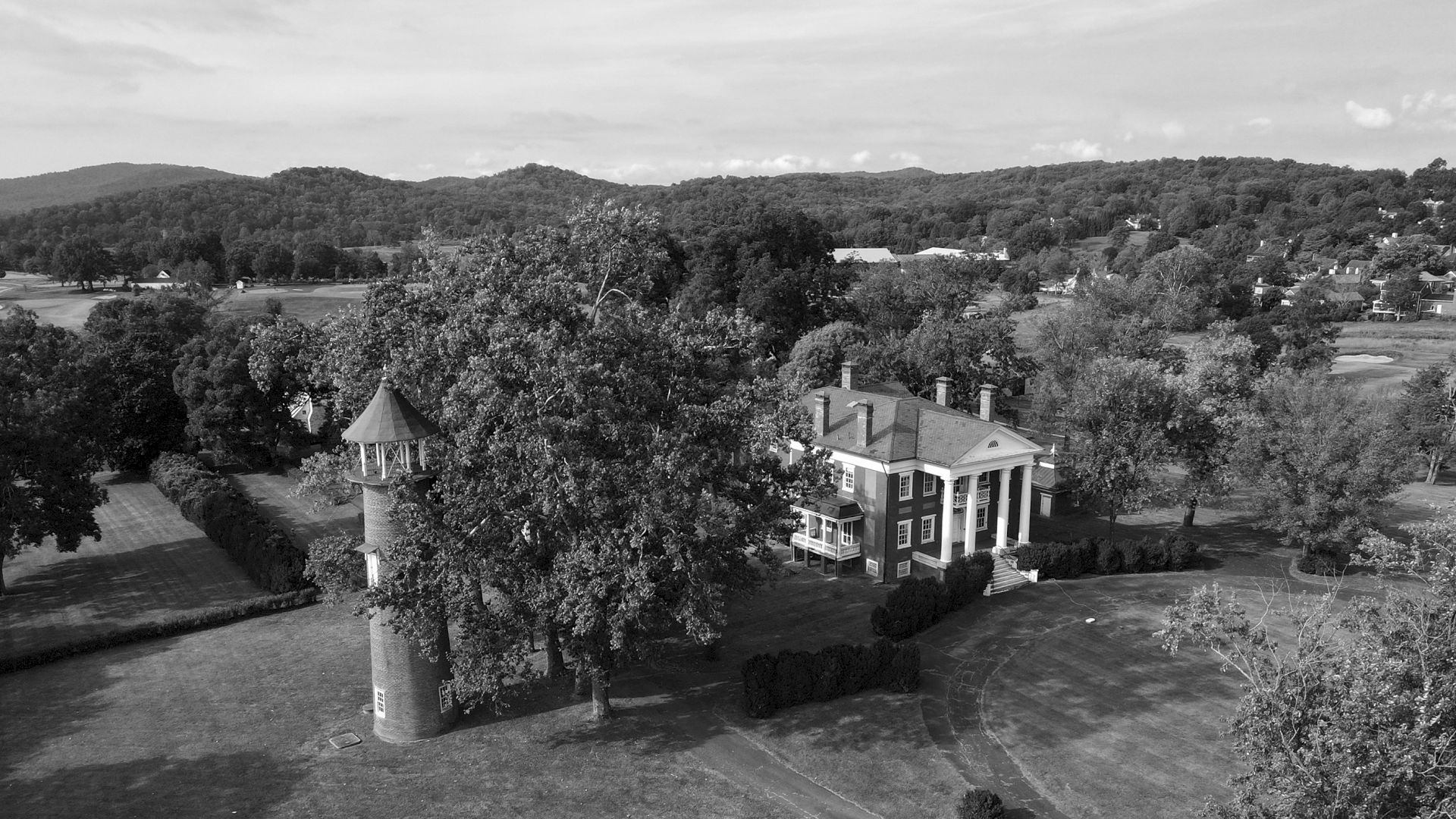An aerial view showcases a historic mansion with columns and a nearby tower, set amidst vast greenery and rolling hills in the background.