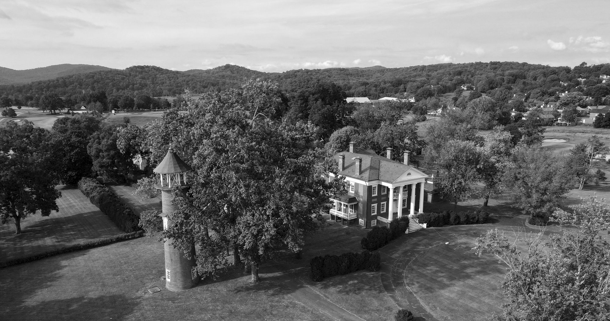 An aerial view showcases a historic mansion with columns and a nearby tower, set amidst vast greenery and rolling hills in the background.