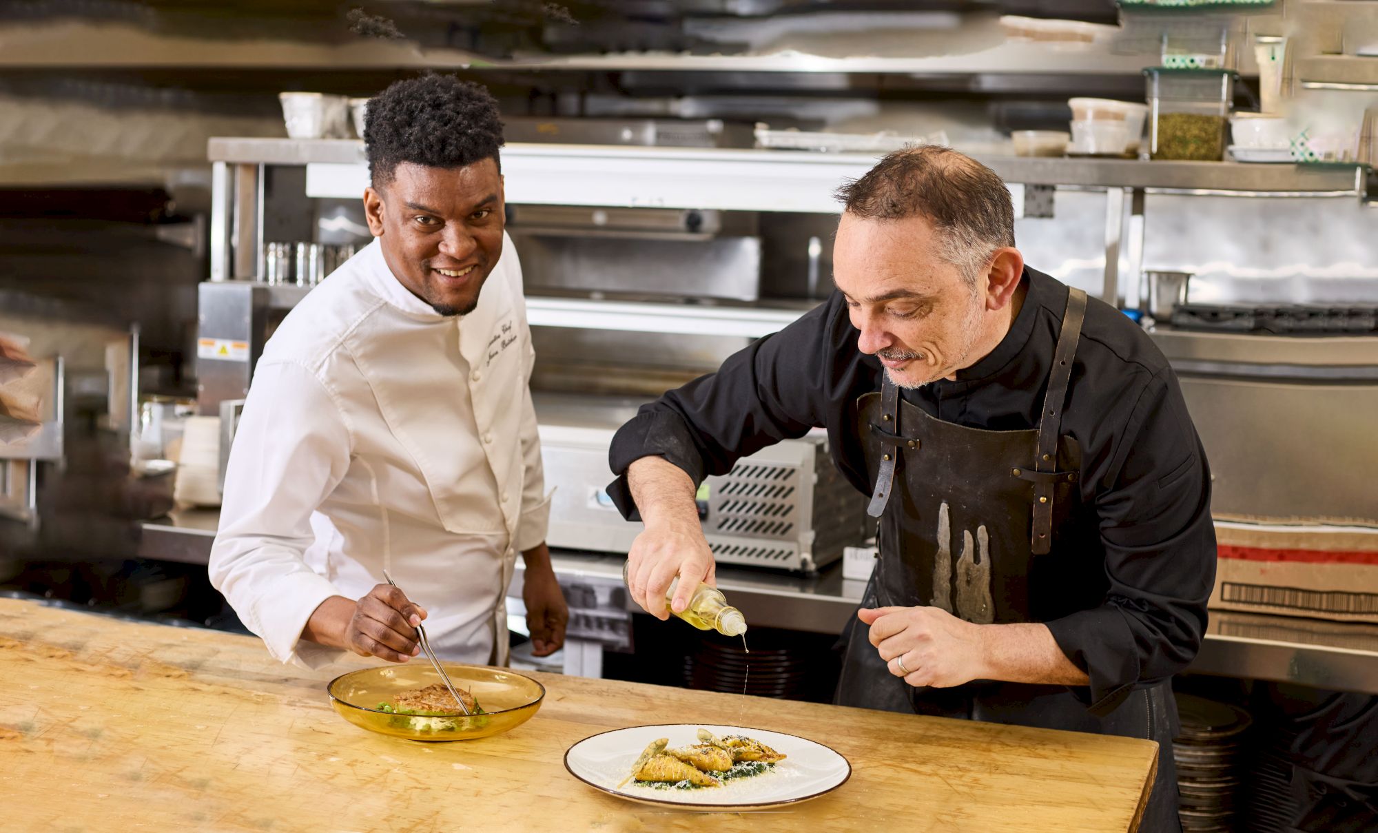 Two chefs in a kitchen garnishing dishes on a wooden counter, focused on presentation and culinary precision.