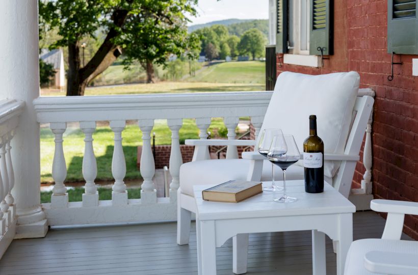 A cozy porch scene with white wicker chair, side table, a book, a wine bottle and two glasses, overlooking a lush green yard.