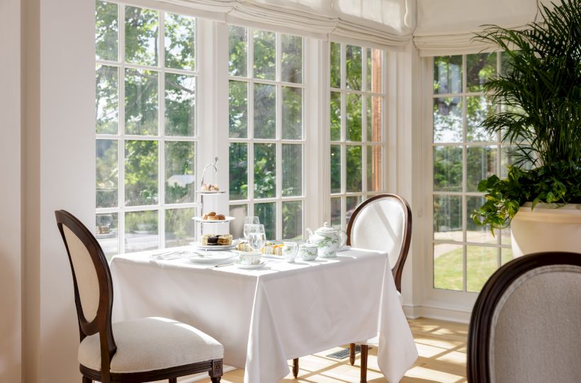 A bright sunlit dining corner with a white-clothed table set for two, elegant chairs, large windows, and a lush green garden view outside.