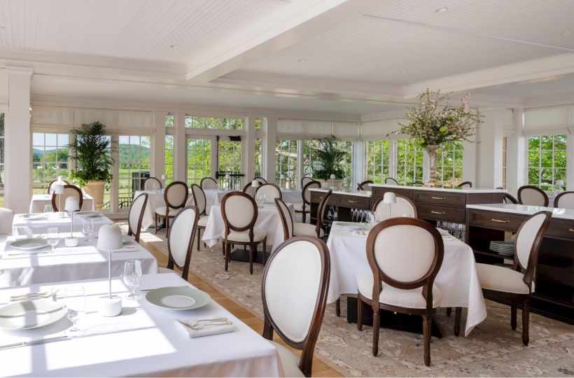 An elegant dining room set for a meal, with white tablecloths, curved chairs, a service counter, and large windows overlooking greenery.
