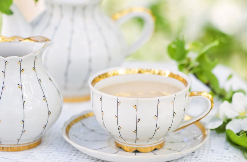 Elegant tea set with a teacup, saucer, and pitcher in white with gold accents, surrounded by greenery and flowers on a lace cloth.