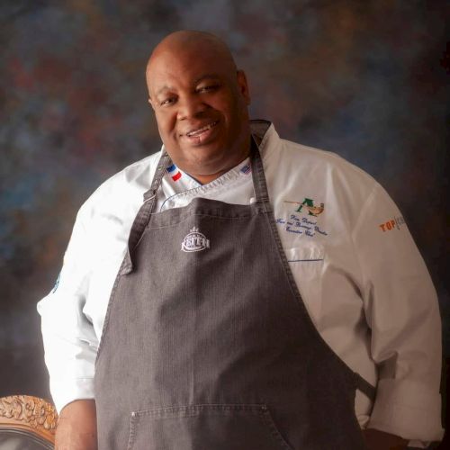 A person wearing a chef's uniform and apron is standing, smiling, with a blurred background, hands resting on a chair.