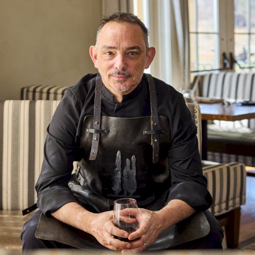 A person in a black chef's outfit sits on a striped couch, holding a drink, with a dining area in the background.