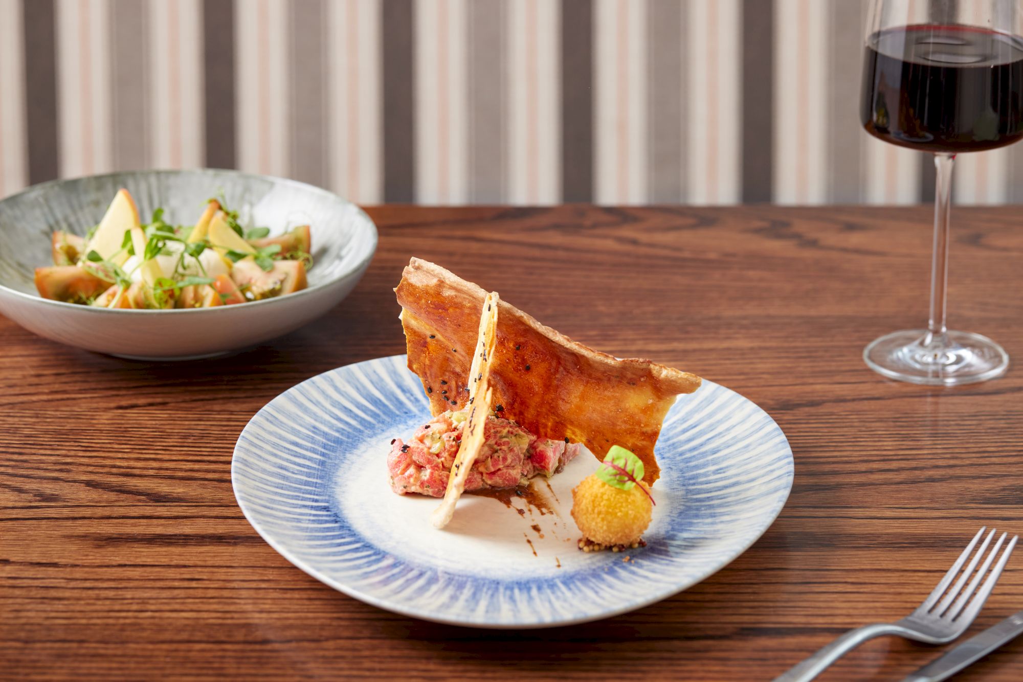 A plate with gourmet food, a side bowl with more food, a fork, and a glass of red wine on a wooden table.