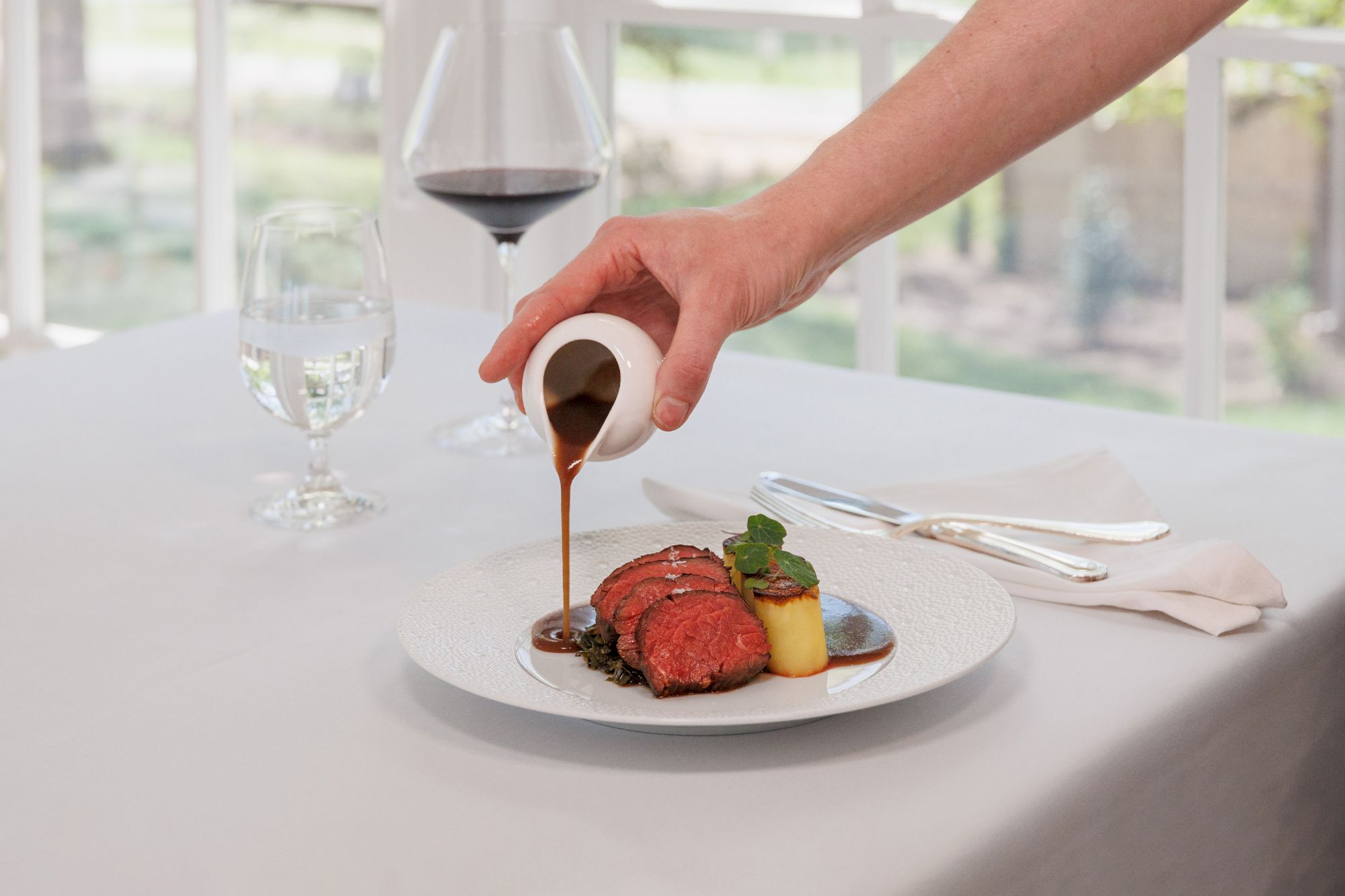 A chef pours a glossy sauce over a plated steak with vegetables on a white table, glass of red wine nearby, elegant dining setting.