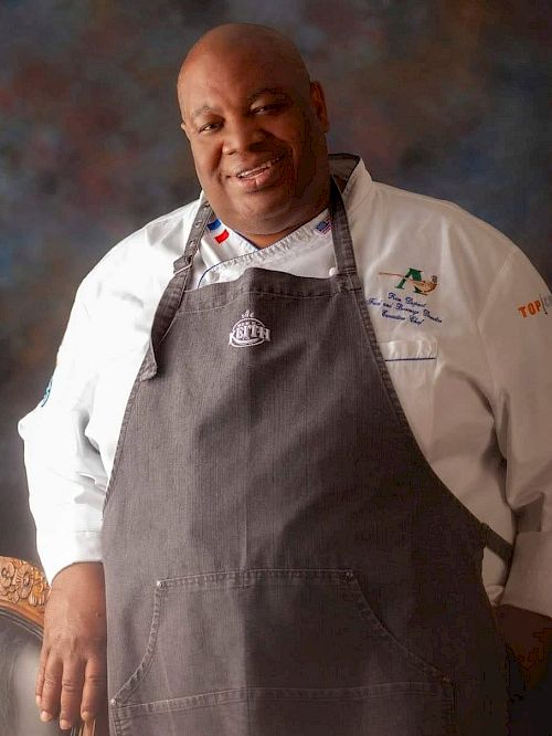 The image shows a person wearing a chef's coat and apron, posing and smiling, with a colorful backdrop behind.