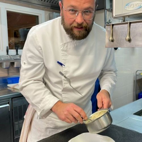 A chef in a kitchen is preparing to plate food, holding a small pot over a white dish while wearing a white chef's coat.