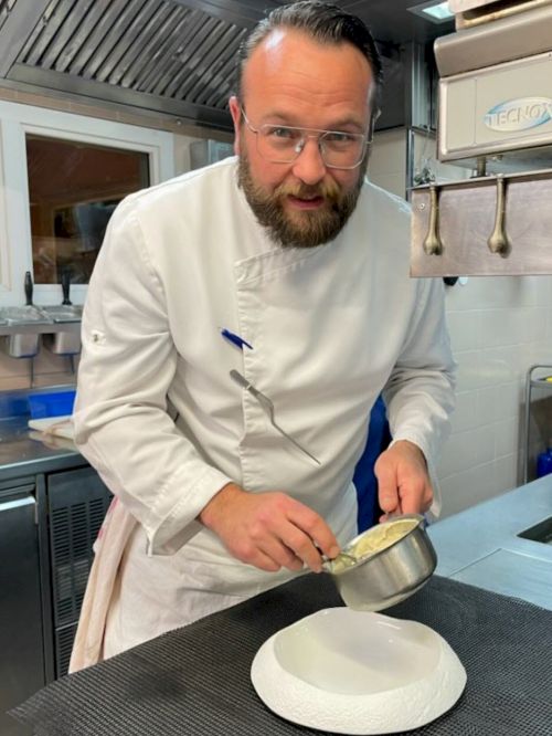A chef in a kitchen is preparing to plate food, holding a small pot over a white dish while wearing a white chef's coat.
