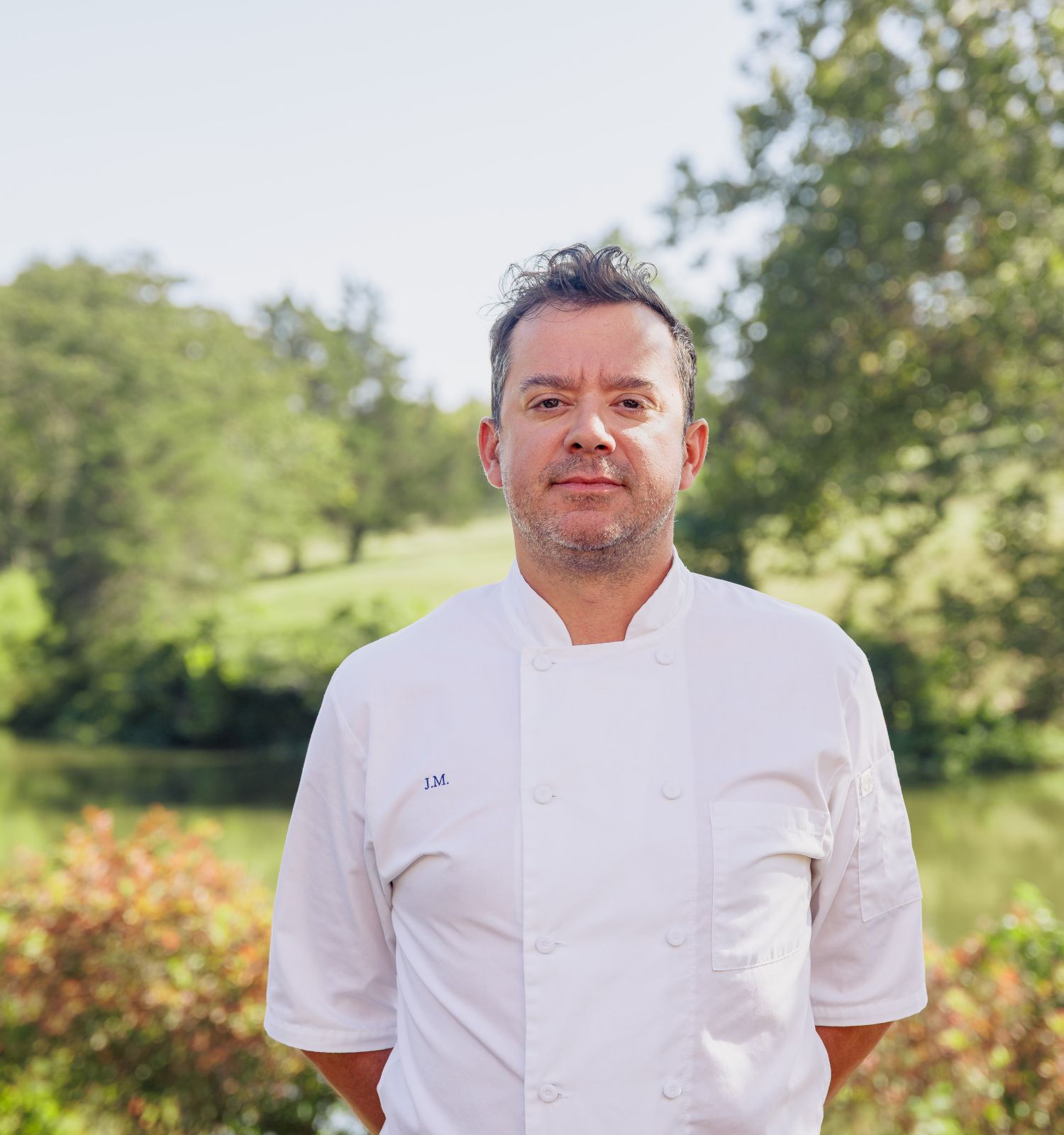 A person in a white chef's coat stands outdoors in front of a scenic, grassy landscape with trees and bushes.
