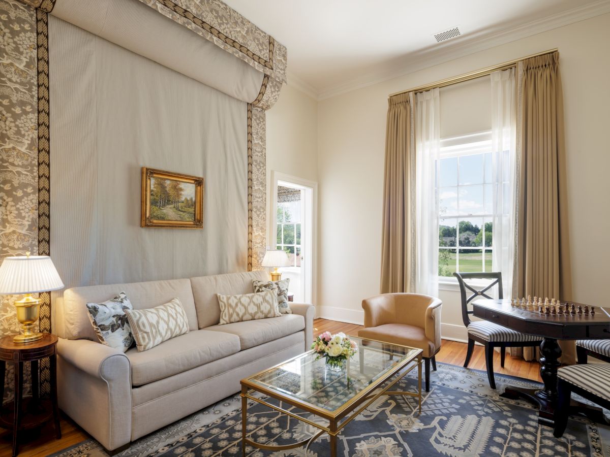A bright, elegant living room with a light sofa, patterned rug, glass coffee table, beige curtains, and a small piano near a window.