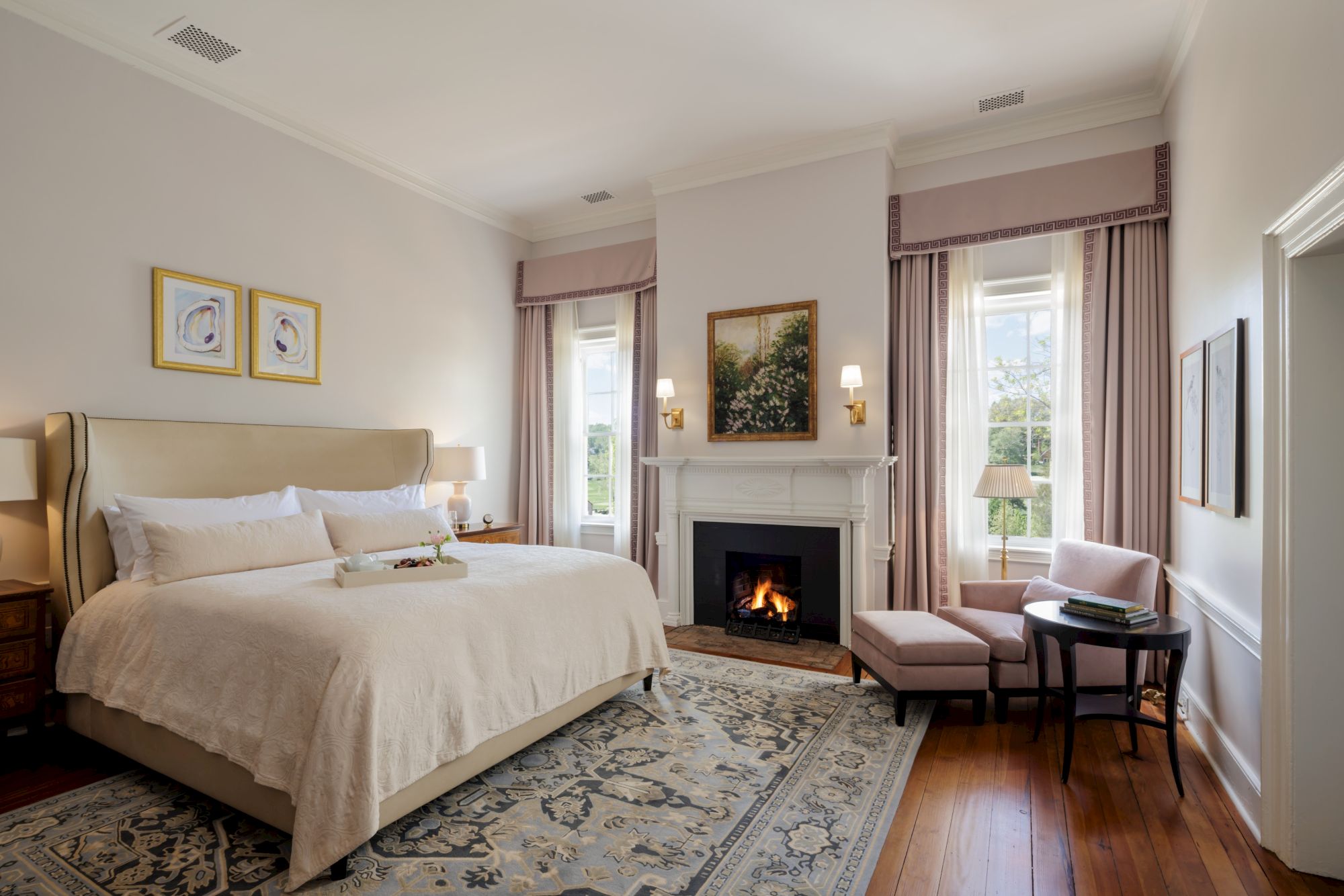 A luxurious hotel room with a plush beige bed, fireplace, armchair by the window, soft pink drapes, and a patterned area rug.