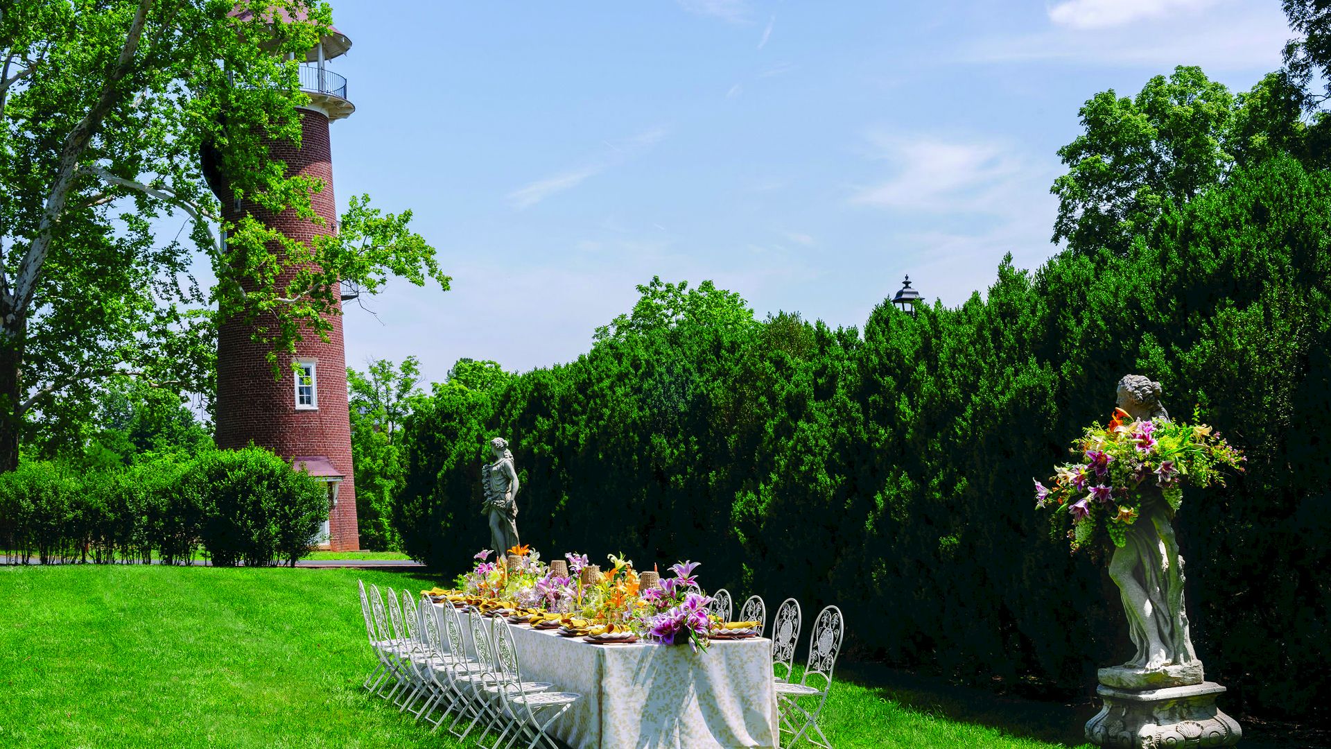 A lush garden party scene with a brick tower, green hedges, flower table, and stone statues.