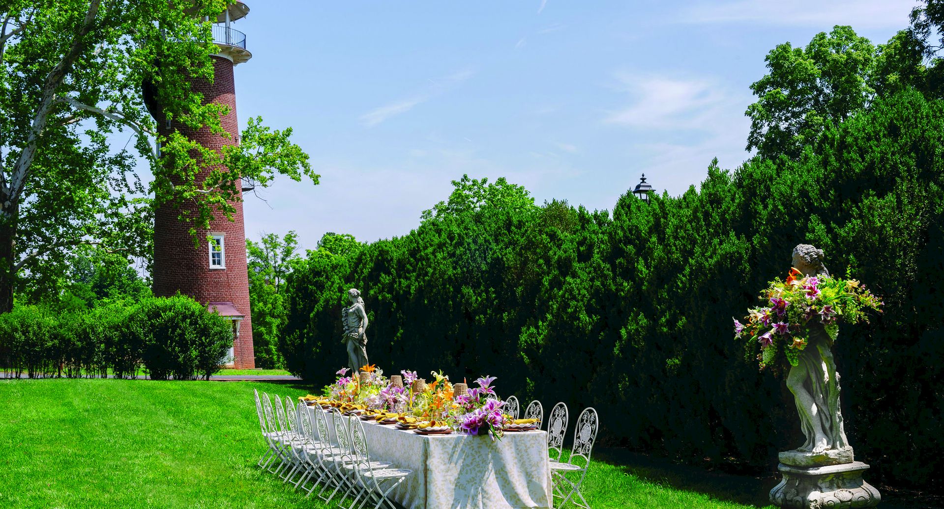 A lush garden party scene with a brick tower, green hedges, flower table, and stone statues.