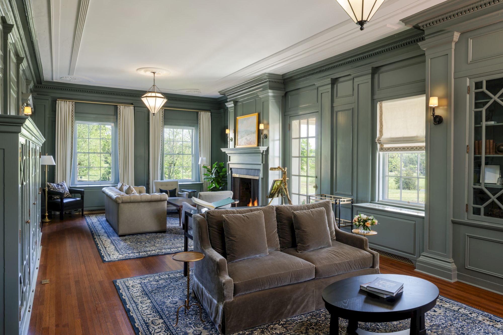A spacious, elegant living room with teal walls, large windows, a stone fireplace, brown sofa, and matching chairs on dark wood floors with a patterned rug, bright and inviting.