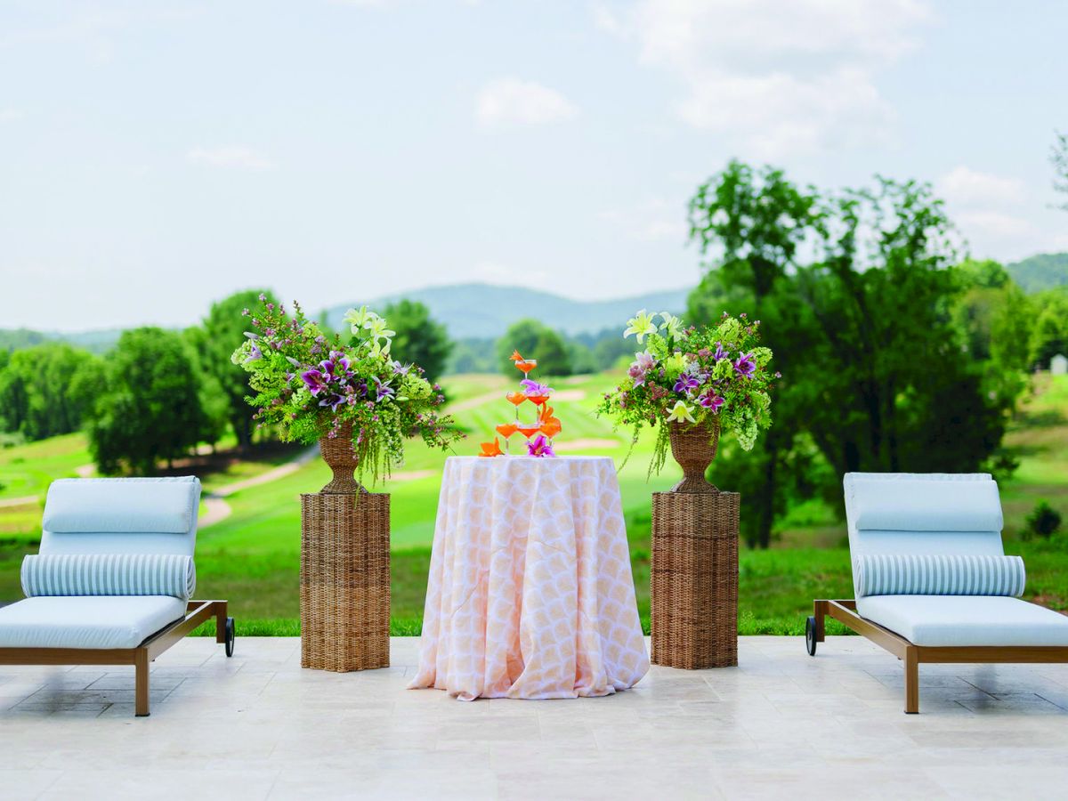 An outdoor patio set with white loungers and a table draped in cloth, surrounded by tall floral centerpieces overlooking a green valley.
