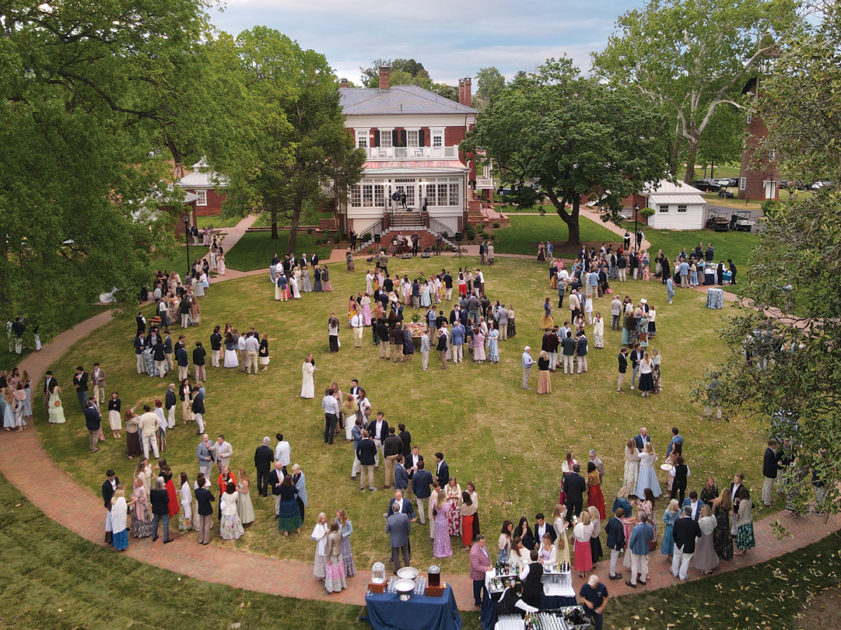 An outdoor wedding reception: many guests in formal attire gather on a lawn before a white-columned brick mansion.