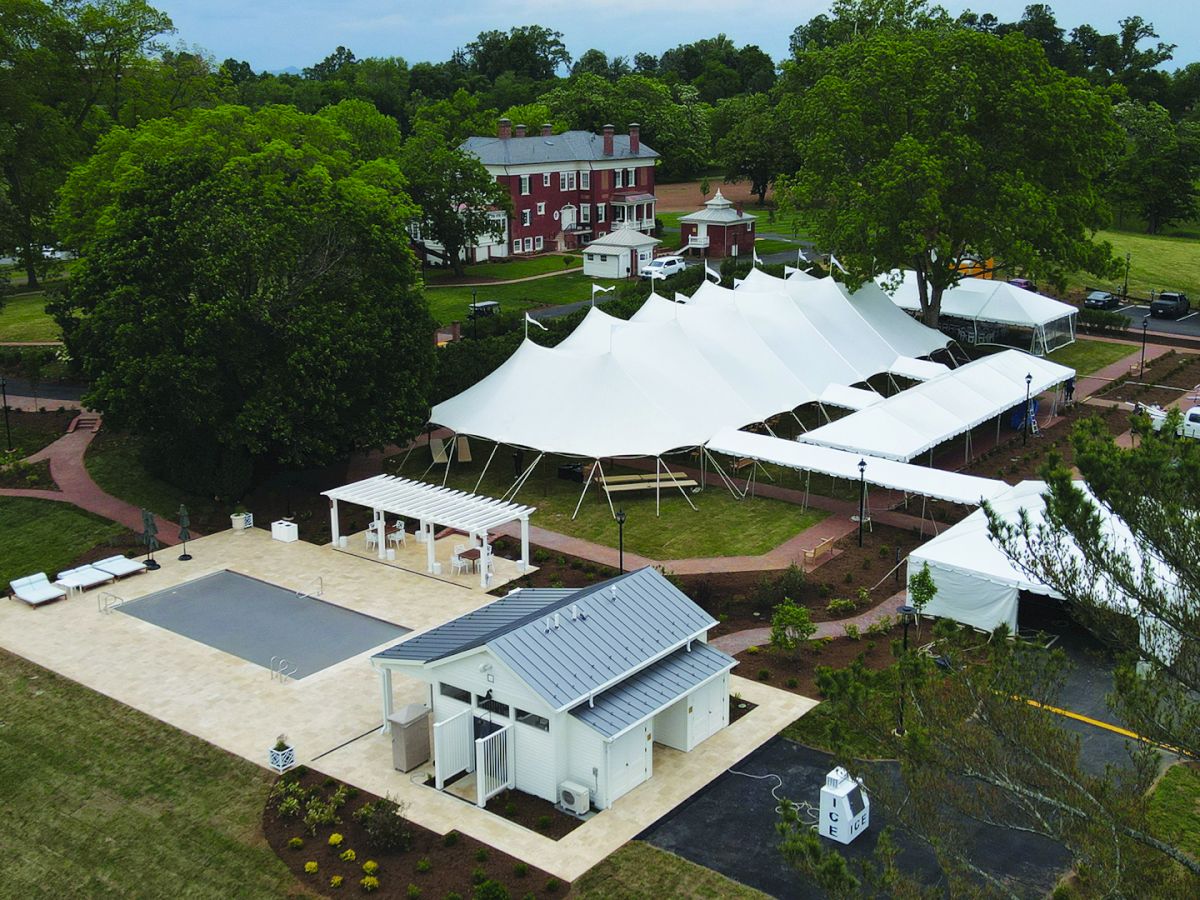 Aerial view of a red mansion estate with a courtyard pool, white event tents, trees, and parked cars on a sunny day.