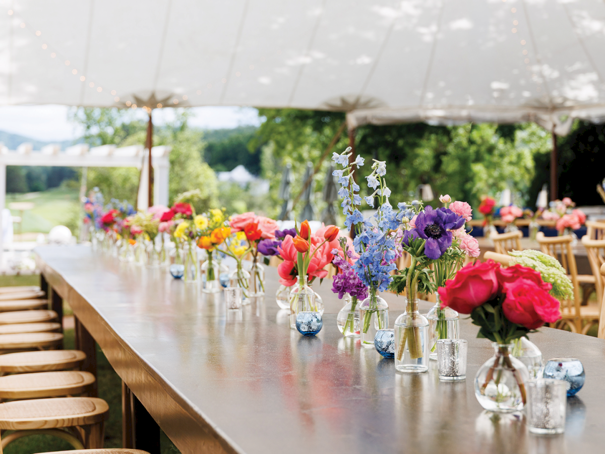 Outdoor event table with colorful bouquets in glass vases, fairy lights overhead, and wooden chairs set for a celebration.