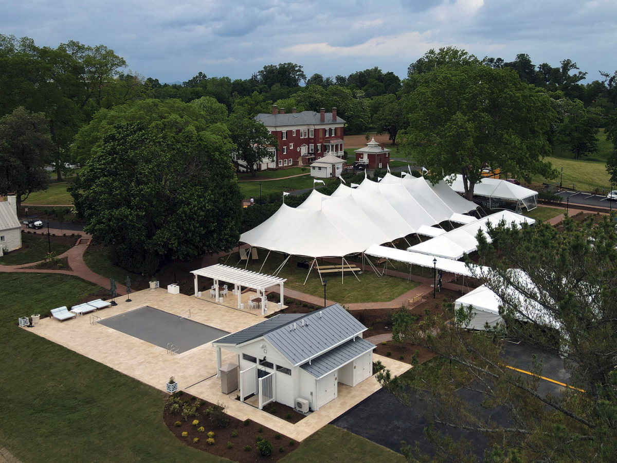 Aerial view of a red manor amid trees, with big white event tents and a courtyard patio below.