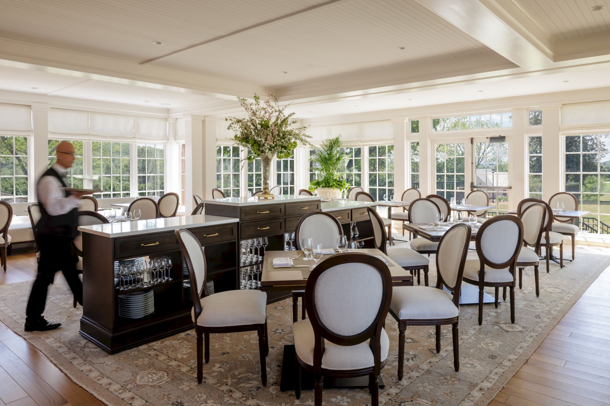 A bright, elegant restaurant or dining room with white chairs around dark wood tables, large windows, and potted plants; a server moves past the bar.