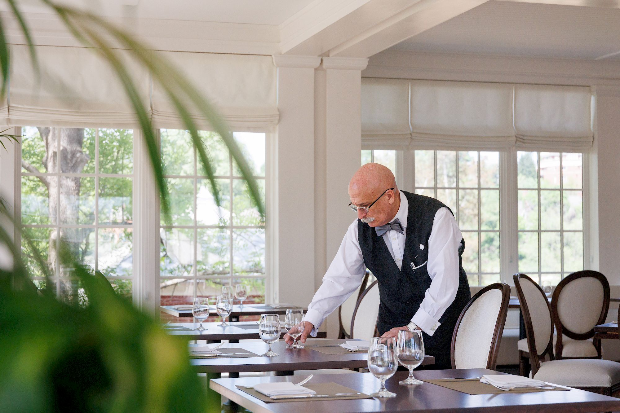 A waiter in a white shirt and vest is tidying a dining table in a bright, elegant restaurant. The room has large windows and empty chairs. End.