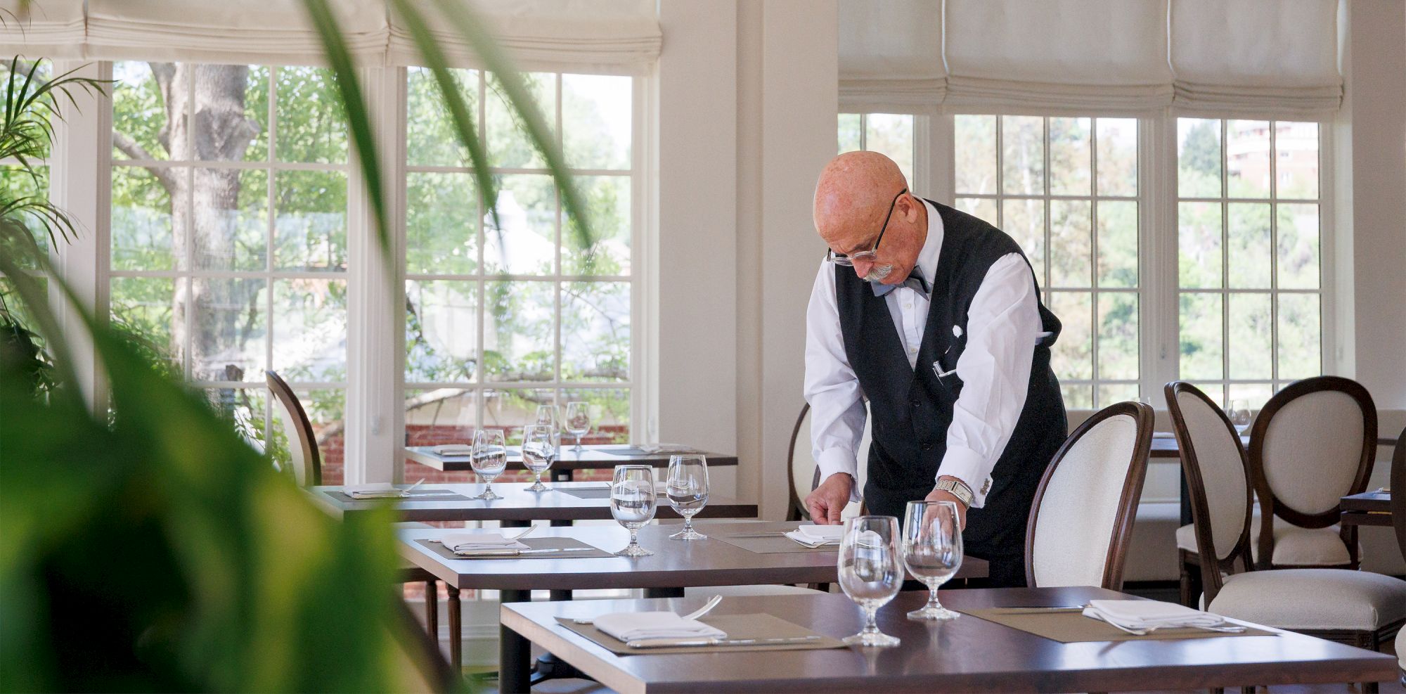 A waiter in a vest is arranging a neatly set dining table in a bright, elegant restaurant with large windows and white chairs.