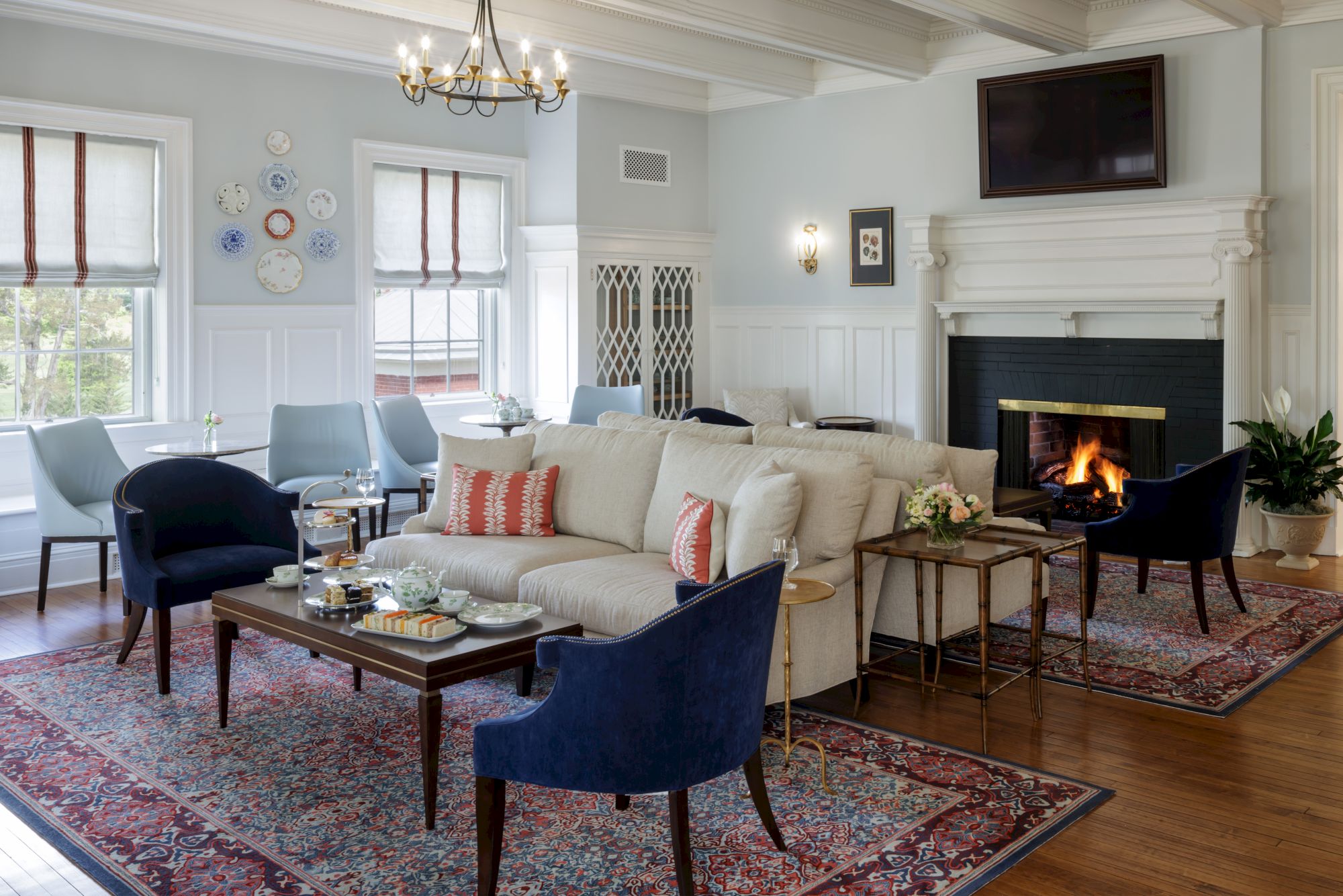 A bright, elegant living room with a white fireplace, cozy beige sofa, navy chairs, patterned area rugs, and a fireplace fire for a warm atmosphere.