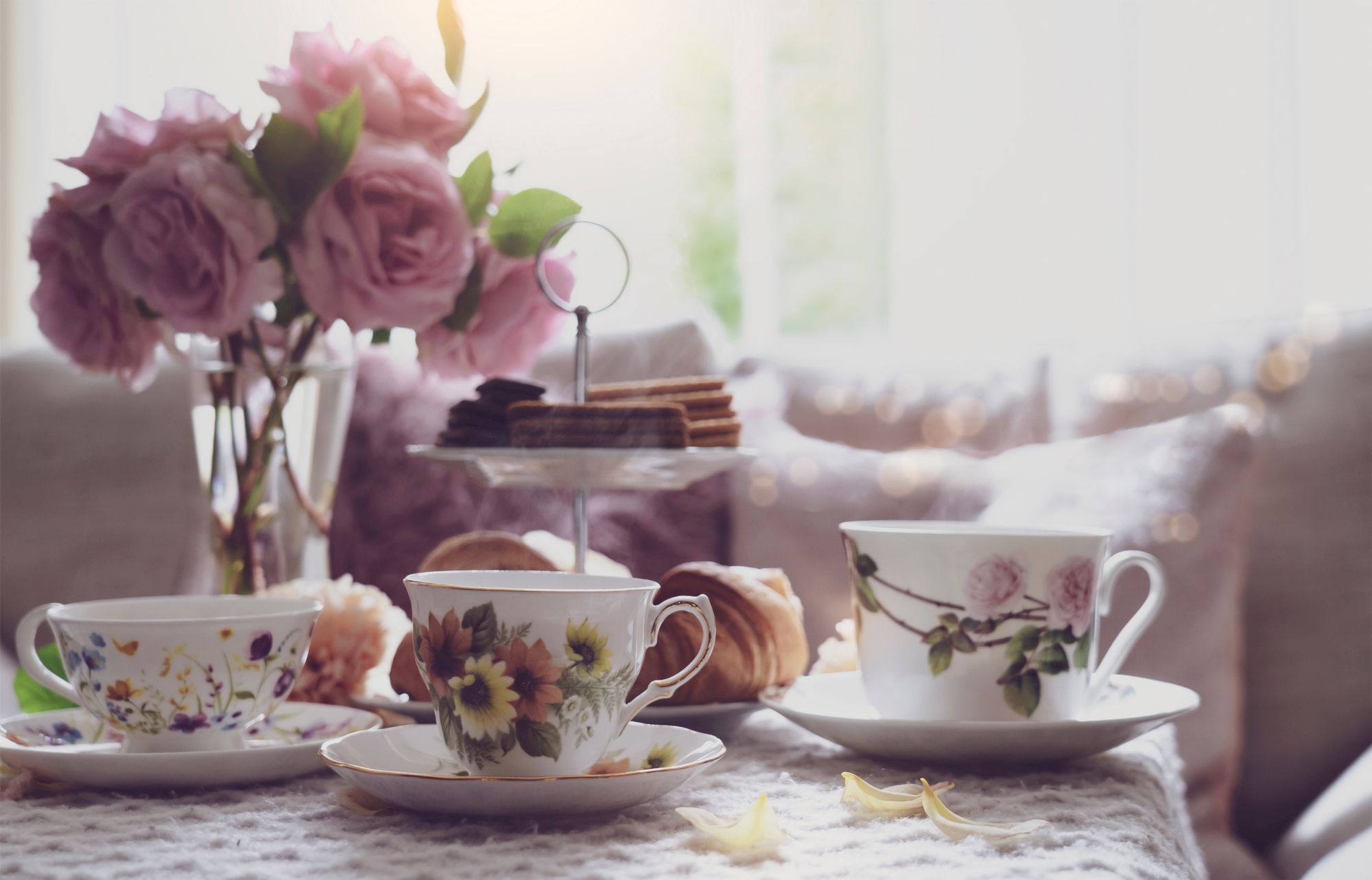 Tea setup with floral teacups, a tiered tray with treats, pink roses in a vase, and a cozy ambiance.