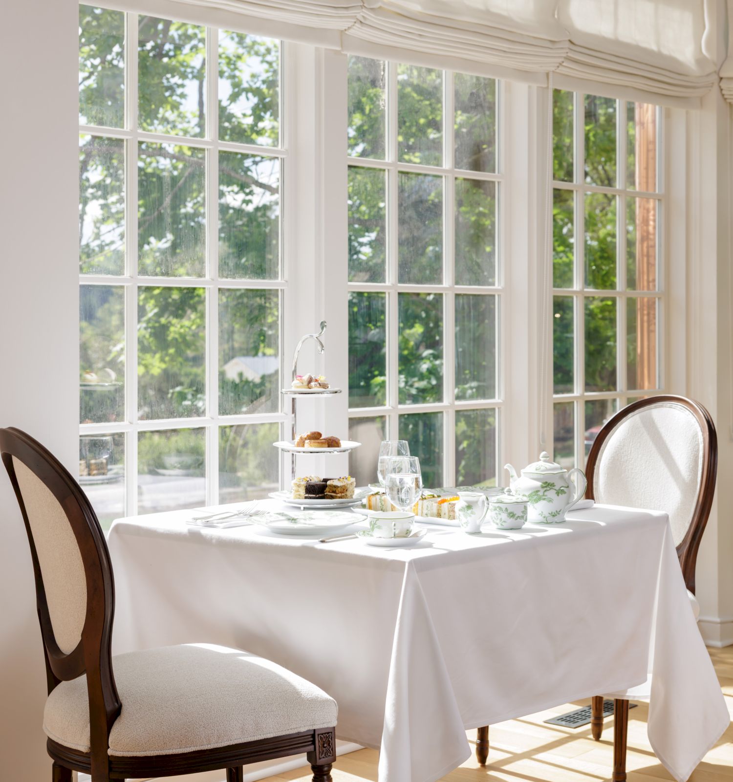A bright dining room by large windows with a white table set for two, elegant chairs, and a plant-filled, sunlit ambiance.