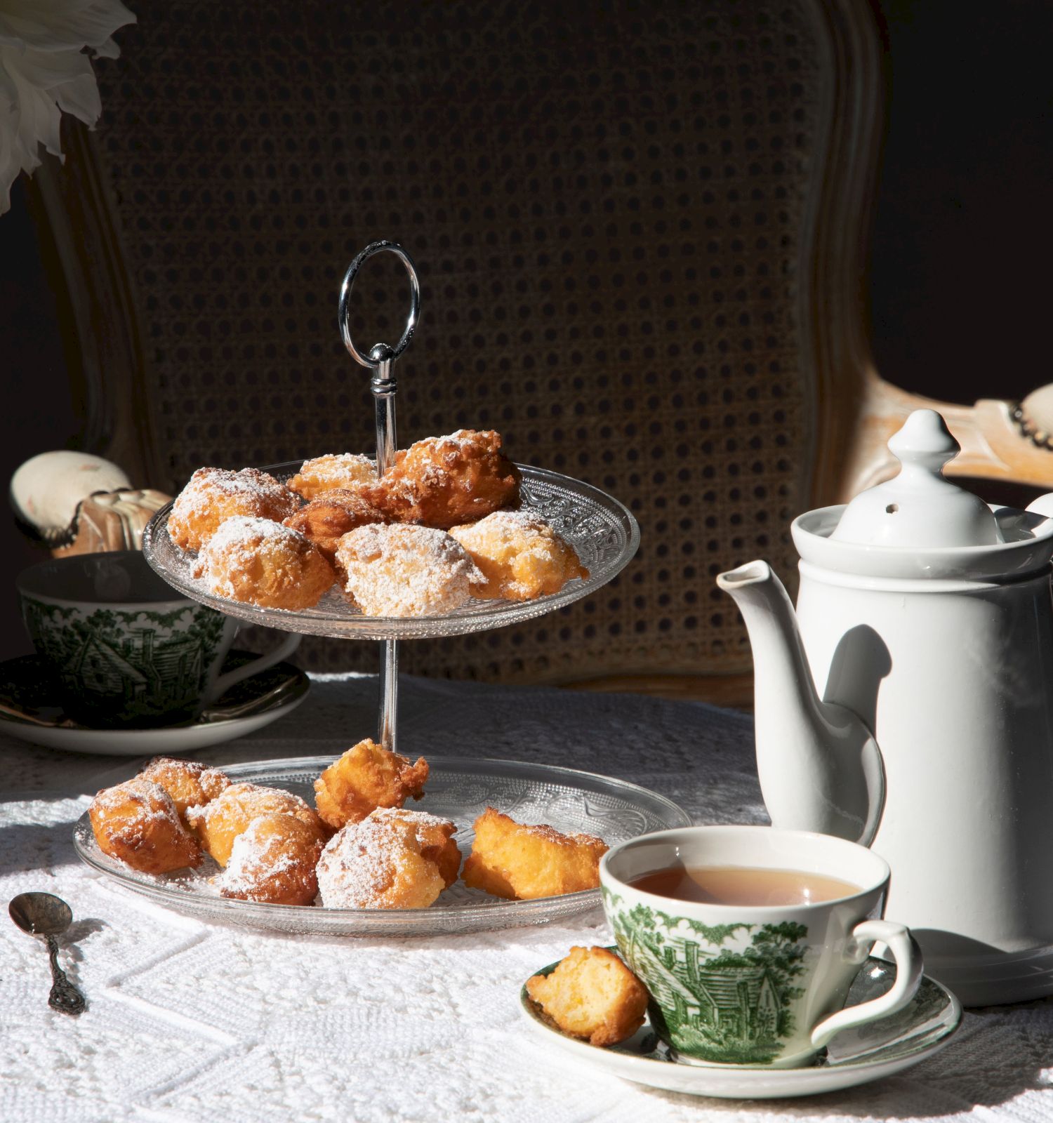 A table set for tea with pastries, a teapot, and teacups on a lace tablecloth, with a chair and flowers in the background.