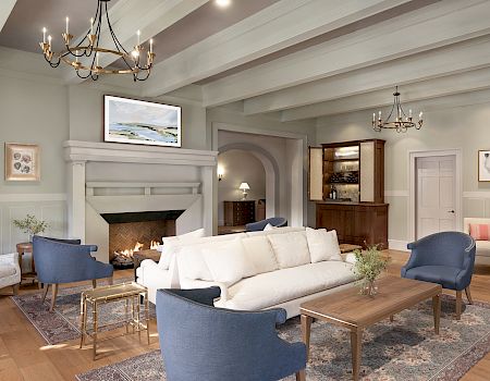 This image shows a cozy living room with a beamed ceiling, elegant furniture, chandeliers, and artwork on the walls.