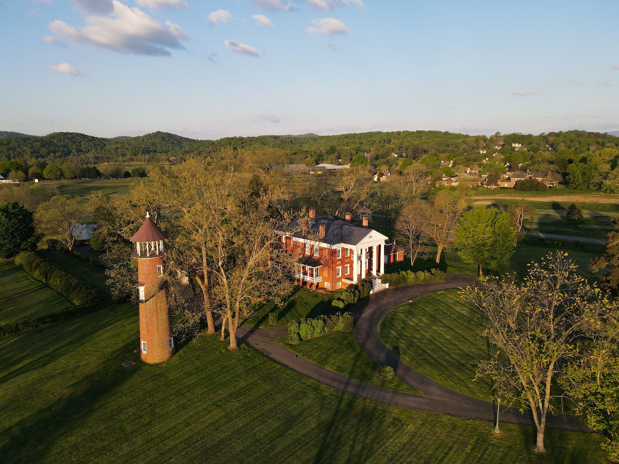 A red-brick house with a wraparound porch sits beside a field, flanked by a tall brick lighthouse-like structure; winding paths circle mature trees on a spacious, sunny campus.